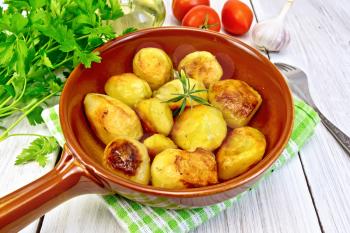 Fried potatoes with rosemary in a ceramic pan on a napkin, garlic, parsley, vegetable oil, tomatoes on a wooden boards background