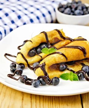 Two pancakes with blueberries, mint and chocolate icing on a white plate, napkin on a wooden boards background