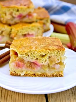 Chunks of sweet cake with rhubarb in a plate, napkin, rhubarb stalks on a wooden boards background