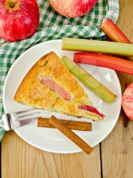 A piece of sweet cake with rhubarb and apples, cinnamon, napkin, fork on a wooden boards background