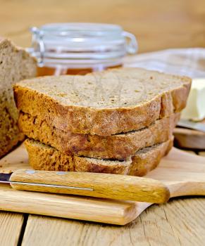 A stack of slices of rye homemade bread with a knife, napkin, loaf of bread, a jar of honey, butter on a wooden boards background