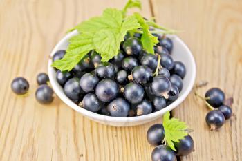 Black currants in a white bowl with green leaves on a wooden boards background