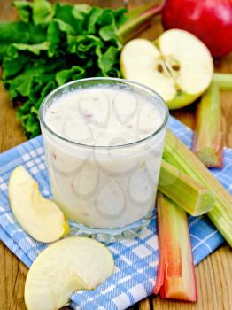 Milkshake in a glassful with leaves and cuttings rhubarb, apple slices on blue napkin on a wooden boards background