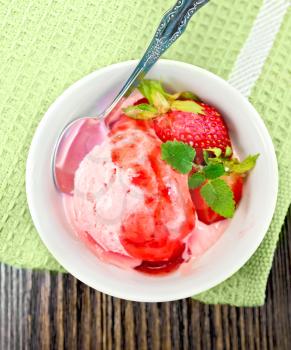 Strawberry ice cream in a white bowl with strawberries, strawberry syrup and a spoon on a napkin on the background of wooden boards on top