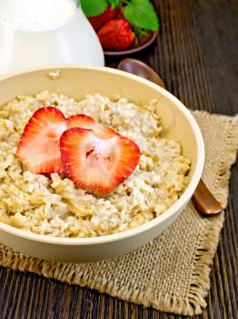 Oatmeal in a bamboo bowl with strawberries on a napkin from a rough burlap, spoon, milk in a glass jar on the background of dark board