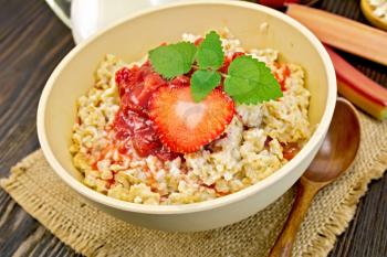 Oatmeal in a bamboo bowl with strawberry and rhubarb sauce on a napkin of burlap, spoon, milk in glass jug, rhubarb on a dark board