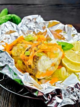 Pike with carrots, leek, basil and lemon slices in a foil on a metal grid, a towel on the background of wooden boards
