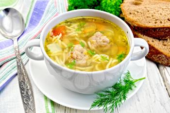 Soup with meatballs, noodles and vegetables in a white bowl and dill on a saucer, bread, napkin and spoon on the background light wooden boards