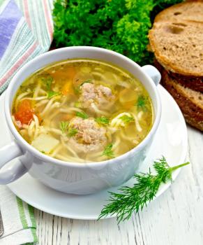 Soup with meatballs, noodles and vegetables in a white cup and saucer, bread, napkin and spoon on the background light wooden boards