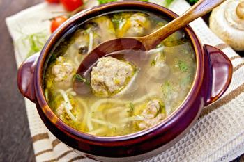Soup with meatballs, noodles and mushrooms in a clay bowl with a wooden spoon on a napkin, parsley, tomatoes and bread on a wooden boards background