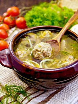 Soup with meatballs, noodles and mushrooms in a clay bowl with a spoon on a napkin, parsley and dill, tomatoes and bread on a wooden boards background