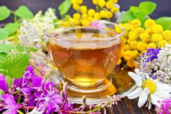 Herbal tea in a glass cup, fresh flowers fireweed, tansy, chamomile, clover, yarrow, meadowsweet, mint leaves on a wooden boards background