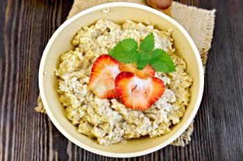 Oatmeal in a yellow cup with strawberries on a napkin of burlap, spoon on dark background board