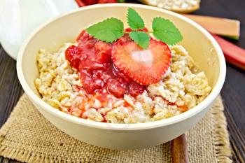 Oatmeal in a yellow cup with strawberry and cherry sauce on a napkin of burlap, spoon, milk in glass jug, rhubarb against the dark boards