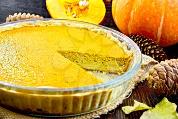 Pumpkin pie in a glass pan on sackcloth, pumpkin, fir cones and maple leaf on the background of dark wood planks