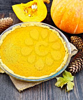 Pumpkin pie in a glass pan, pumpkin, cones and maple leaf on the background of dark wood planks
