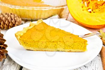 A piece of pumpkin pie with a fork in the white plate, pumpkin and fir cones on the background light wooden boards
