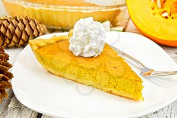 A piece of pumpkin pie with whipped cream, a fork in the white plate, pumpkin and fir cones on the background light wooden boards
