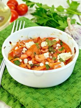Shrimp and tomatoes baked with feta cheese in a white bowl on a green towel, parsley and fork on a wooden boards background