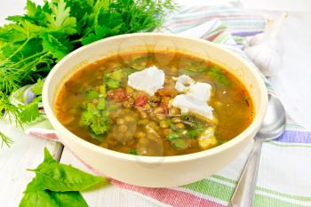 Lentil soup with spinach, tomatoes and feta cheese in a yellow bowl, spoon on a kitchen towel, parsley and spinach on the background light wooden boards