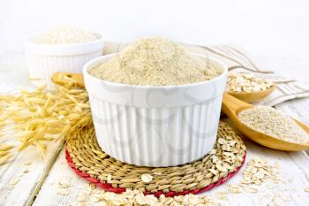Oat flour in white bowl, oatmeal and bran in a spoon, stalks of oats, napkin against the background of wooden boards