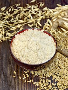 Oat flour in a bowl, bag with oat grains and stalks of oats on the background of wooden boards