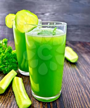 Cucumber juice in two tall glass with slices of cucumber, cucumber and parsley on a dark wooden board
