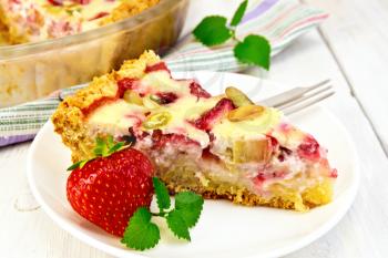Piece of pie with strawberries, rhubarb and cream sauce, fork, strawberries, mint in white plate, a glass pan with pie on a napkin on a wooden boards background