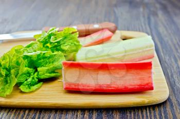 Rhubarb stalks with green leaves and a knife on a small planch on a dark wooden board