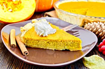 A piece of pumpkin pie with whipped cream in a brown plate with a fork and cinnamon, pumpkin on a dark wooden board
