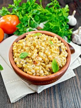 Barley porridge in a clay bowl with basil on a napkin, tomatoes, parsley and garlic on a wooden boards background