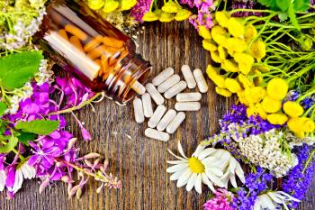 Capsules in brown jar open and on table, fresh flowers fireweed, tansy, chamomile, clover, yarrow, meadowsweet, mint leaves on background of wooden boards top