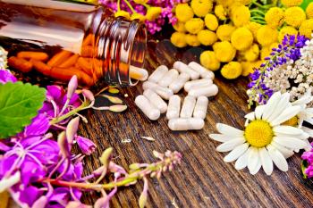 Capsules in a brown jar and on the table, flowers of fireweed, tansy, chamomile, clover, yarrow, meadowsweet, mint leaves on a background of wooden boards