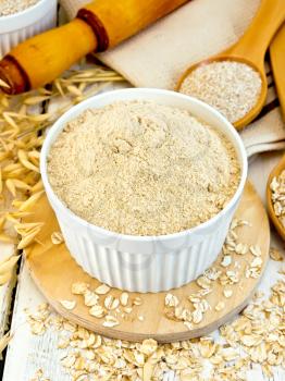Oat flour in white bowl, oatmeal and bran in a spoon, oat stalks, a rolling pin and a napkin on a wooden boards background
