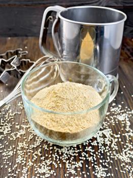 Sesame flour in a glass cup with strainer and mixer, sesame seeds on a wooden boards background