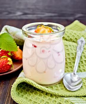 Yogurt with strawberries in a glass jar, spoon on a napkin, strawberry and mint and a plate on a wooden boards background