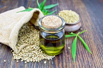 Hemp oil in a glass jar with flour in a clay bowl and grain in a bag, cannabis leaves and stalks on a wooden boards background