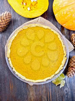 Pumpkin pie in a glass pan on sackcloth, cones and maple leaf against the dark wooden boards on top