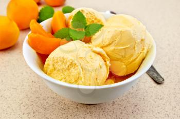 Apricot ice cream in a white bowl with slices of fruit and mint, a spoon on a background granite table