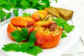 Tomatoes stuffed with meat and steamed wheat bulgur, parsley in a white plate, napkin, fork and bread on the background light wooden boards