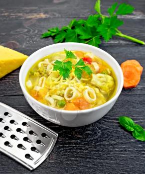 Minestrone soup with meat, celery, tomatoes, zucchini and cabbage, green peas, carrots and pasta in a white bowl, cheese, grater and parsley on a background black wooden plank