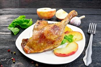 Roasted duck leg with apple, potatoes in a white plate, basil, garlic and fork on black background wooden plank