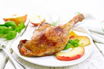 Roasted duck leg with apple, potatoes in a white plate, basil, fork and kitchen towel on a background of wooden boards