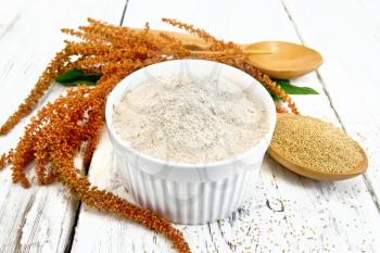 Amaranth flour in white bowl, spoons with grain, brown flower with green leaves on a background of wooden boards