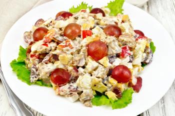 Salad of meat, salty soft feta cheese, sweet pepper, egg and grape with mayonnaise on lettuce the plate, fork on the background light wooden boards