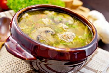Soup with meatballs, noodles and champignon in a clay bowl, spoon on the towel, parsley, tomatoes, mushrooms and bread on dark background wooden plank