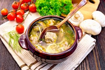 Soup with meatballs, noodles and champignon in a clay bowl with spoon on a towel, parsley, mushrooms, tomatoes and bread on a wooden boards background