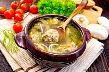 Soup with meatballs, noodles and champignon in a clay bowl with spoon on a napkin, parsley, mushrooms, tomatoes and bread on a dark wooden board