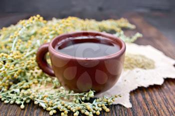 Herbal tea in a brown clay cup, twigs gray sagebrush, wormwood dried on a paper on the background of a wooden tabletop