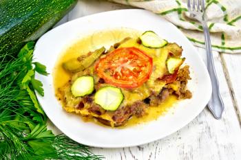 Casserole of minced meat, tomatoes and zucchini in a plate, napkin and fork on a wooden plank background
